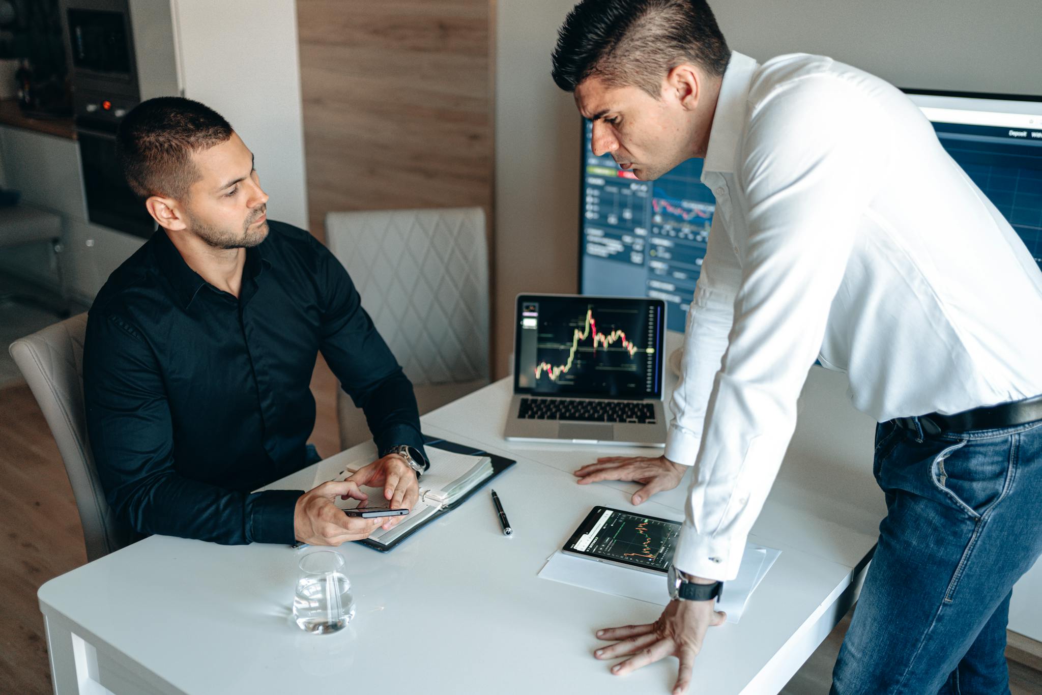 Two businessmen in an office analyzing stock market charts on digital devices with focused discussion.